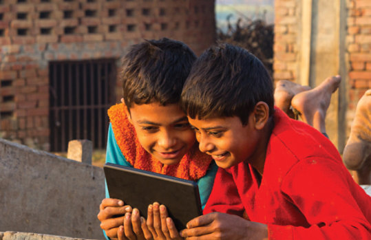 Two children lying on a wooden cart outdoors, smiling while looking at a tablet in a rural village setting.